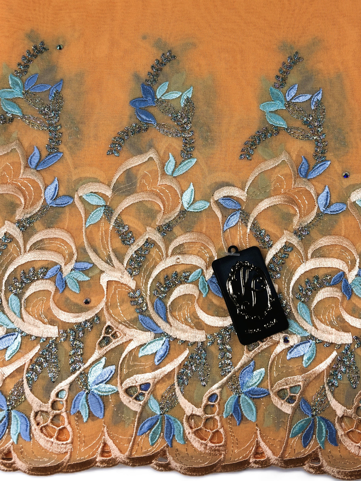 A close-up of an embroidered fabric with a peach-orange base, featuring beige and blue floral patterns with metallic silver beadwork. A black fabric tag is placed on the design.